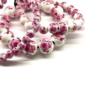 May include: A string of white ceramic beads with pink floral designs. The beads are strung together with a black cord.