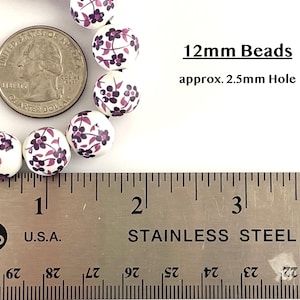 Pu&ograve; includere: Perline bianche da 12 mm con un motivo floreale viola e rosa. Le perline sono mostrate accanto a una moneta per il confronto delle dimensioni e un righello. Il testo sull'immagine recita "12mm Beads approx. 2.5mm Hole."