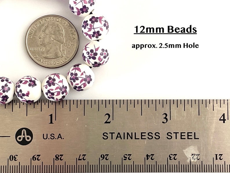 Pu&ograve; includere: Perline bianche da 12 mm con un motivo floreale viola e rosa. Le perline sono mostrate accanto a una moneta per il confronto delle dimensioni e un righello. Il testo sull'immagine recita "12mm Beads approx. 2.5mm Hole."