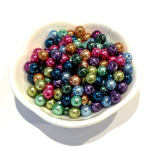 May include: A collection of small, round, multicolored faux pearl beads in shades of pink, blue, green, purple, and bronze, contained within a white, flower-shaped ceramic dish. Each bead features a central hole for threading.