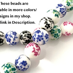 Two strands of floral ceramic porcelain beads lie twisted on a white surface.  The beads are white with black blue green and pink flowers.  Text in the upper left corner says that there is a link in the Description to beads in additional colors