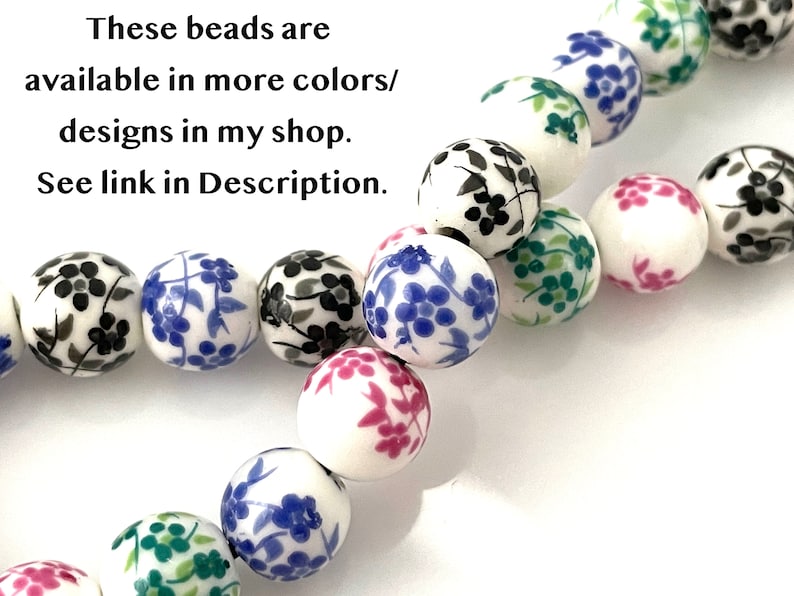 Two strands of floral ceramic porcelain beads lie twisted on a white surface.  The beads are white with black blue green and pink flowers.  Text in the upper left corner says that there is a link in the Description to beads in additional colors
