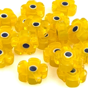 May include: A group of yellow glass flower beads with blue and white centers. The beads are scattered on a white surface.