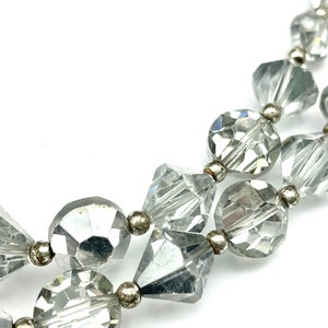 May include: Close-up of a clear, faceted bead necklace with silver-toned metal accents. The beads are various shapes, including round, square, and diamond-like, creating a sparkling effect. The necklace is displayed against a white background.