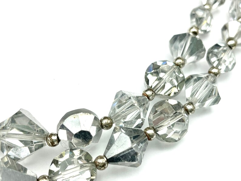 May include: Close-up of a clear, faceted bead necklace with silver-toned metal accents. The beads are various shapes, including round, square, and diamond-like, creating a sparkling effect. The necklace is displayed against a white background.