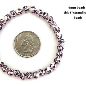 6mm porcelain ceramic beads with a purple floral pattern on a white background are strung in a loop that lies on a flat white background next to a quarter to show scale.