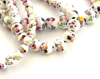 May include: White ceramic beads with a floral pattern in shades of pink, purple, and yellow. The beads are strung together to form a necklace or bracelet.