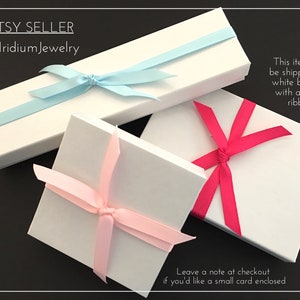 May include: Three white gift boxes with different colored ribbons. The boxes are tied with a light blue ribbon, a pink ribbon, and a hot pink ribbon. The text "This item will be shipped in a white box tied with a fabric ribbon" is printed on the right side of the image. The text "Leave a note at checkout if you'd like a small card enclosed" is printed on the bottom of the image.