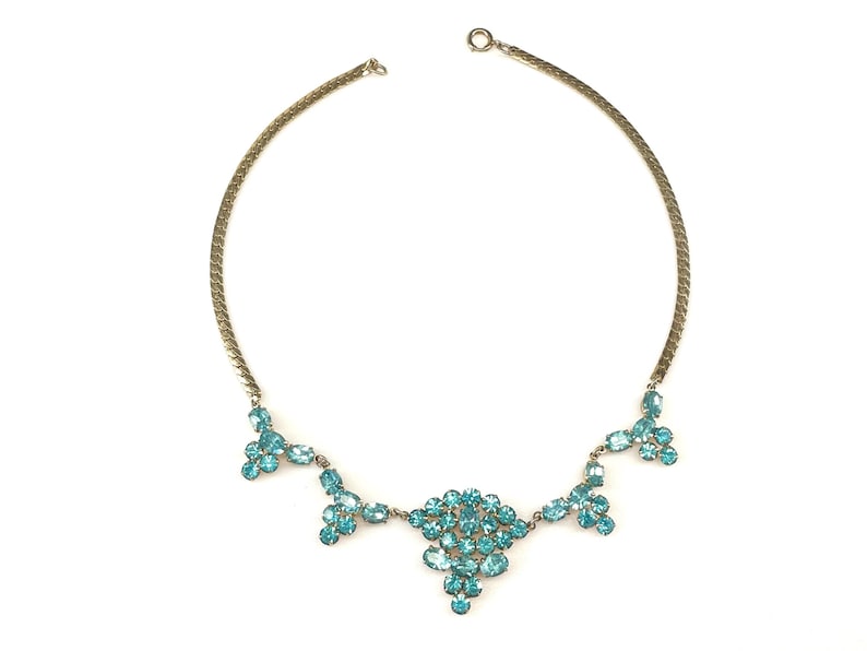 May include: A gold chain necklace with a cluster of turquoise blue rhinestones in the center and smaller clusters on either side.