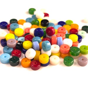 May include: A collection of colorful, round beads in various shades of blue, green, yellow, red, orange, and white. The beads have a donut shape with a hole in the center. The beads are scattered on a white surface.
