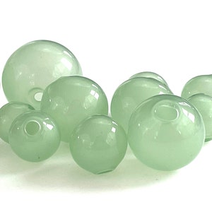 May include: A group of pale green glass beads of various sizes. The beads are smooth and round, with a slight sheen.