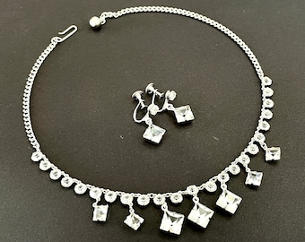Può includere: Una collana in metallo color argento con una catena di piccoli cristalli rotondi trasparenti e pendenti in cristallo quadrato più grandi, abbinata a orecchini a vite coordinati con un design a goccia in cristallo quadrato.