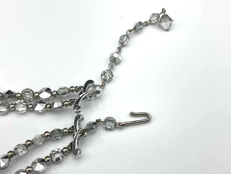 May include: A silver-toned necklace with multiple strands of faceted, clear beads. The necklace features decorative clasps and a hook closure. The beads are various shapes and sizes, reflecting light. The jewelry is set against a white background.