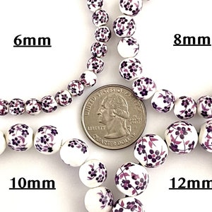 Pu&ograve; includere: Perline di ceramica bianca con un motivo floreale nei toni del viola. Le perline sono mostrate nelle misure di 6 mm, 8 mm, 10 mm e 12 mm. &Egrave; inclusa una moneta da un quarto di dollaro USA per il confronto delle dimensioni. Il testo "6mm", "8mm", "10mm" e "12mm" &egrave; visualizzato.