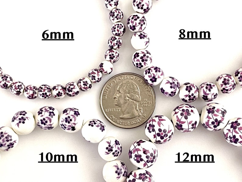 Pu&ograve; includere: Perline di ceramica bianca con un motivo floreale nei toni del viola. Le perline sono mostrate nelle misure di 6 mm, 8 mm, 10 mm e 12 mm. &Egrave; inclusa una moneta da un quarto di dollaro USA per il confronto delle dimensioni. Il testo "6mm", "8mm", "10mm" e "12mm" &egrave; visualizzato.