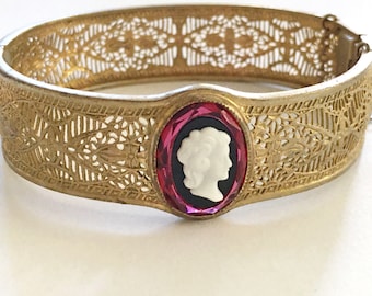 May include: A gold filigree bangle bracelet with a cameo featuring a white profile of a woman set in a red rhinestone. The bangle is open and has a delicate design.