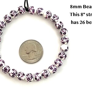 A circular strand of 8mm white porcelain ceramic beads with a purple floral print lie flat on a white surface next to a US quarter to show size.  Text in the upper right hand corner indicates that an 8 inch strand of 8mm beads includes 26 beads