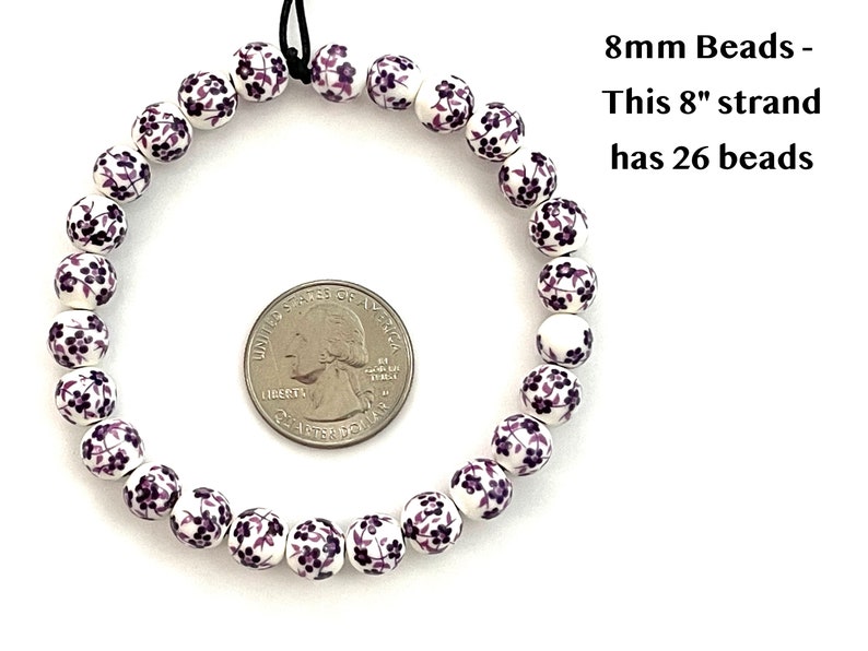 A circular strand of 8mm white porcelain ceramic beads with a purple floral print lie flat on a white surface next to a US quarter to show size.  Text in the upper right hand corner indicates that an 8 inch strand of 8mm beads includes 26 beads