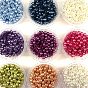 May include: Nine clear plastic containers filled with round, smooth, glossy beads in various colors including light blue, dark blue, cream, purple, magenta, lime green, copper, and white, arranged in a three-by-three grid pattern.