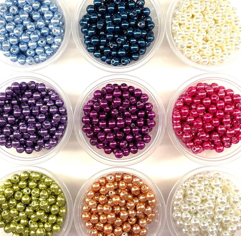 May include: Nine clear plastic containers filled with round, smooth, glossy beads in various colors including light blue, dark blue, cream, purple, magenta, lime green, copper, and white, arranged in a three-by-three grid pattern.