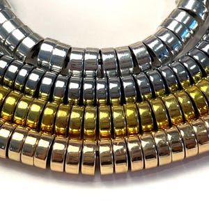 May include: Close-up of a necklace featuring multiple strands of metallic rings. The necklace has silver and gold-toned sections, creating a layered effect. The rings are closely linked, forming a flexible, textured surface.