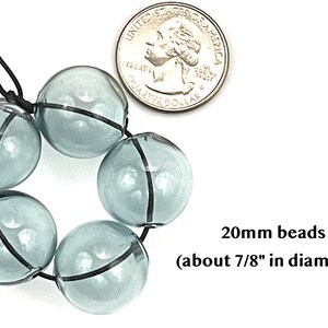 20mm hollow gray blue glass beads sit on a flat white surface next to a US quarter to show size.  These lampwork bubble beads have two holes and are grey with a light blue cast.
