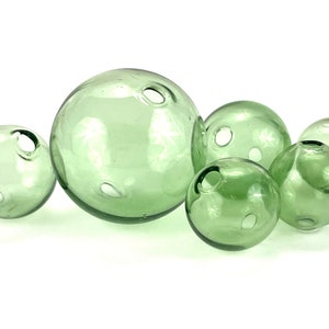 May include: A group of six green glass floats with holes in them. The floats are various sizes and are arranged in a cluster on a white background.