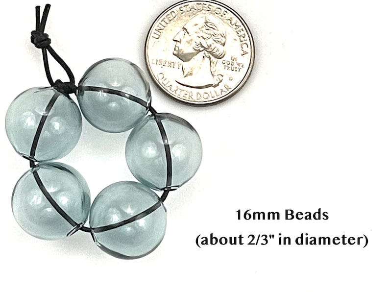 16mm hollow gray blue glass beads sit on a flat white surface next to a US quarter to show size.  These lampwork bubble beads have two holes and are grey with a light blue cast.