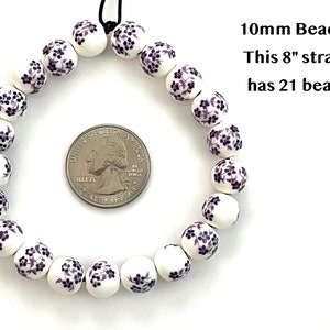 A circular strand of 10mm white porcelain ceramic beads with a purple floral print lie flat on a white surface next to a US quarter to show size.  Text in the upper right hand corner indicates that an 8 inch strand of 10mm beads includes 21 beads