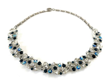 May include: A silver-tone metal necklace featuring a series of articulated floral-shaped links. The design is accented with round dark blue and iridescent light blue rhinestones set within the textured silver petals and along the chain.