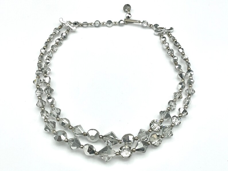 May include: A two-strand silver-toned necklace with clear, faceted beads. The necklace features a variety of bead shapes, including round, faceted, and geometric. The clasp is a simple hook and eye closure.