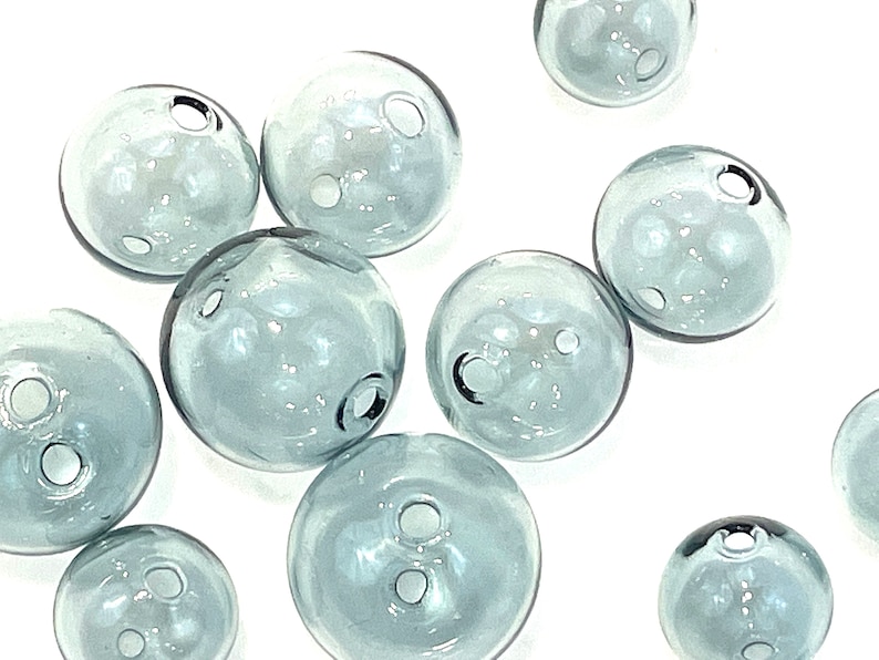 Top view of hollow gray blue glass beads sitting on a flat white surface.  These lampwork bubble beads have two holes and are shown in sizes  of 14mm 16mm and 20mm in diameter.  These are grey with a light blue cast.