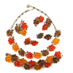 May include: A jewelry set consisting of a necklace, a bracelet, and three pairs of clip-on earrings. Each piece features translucent resin leaves in shades of orange, red, and brown, accented with small clear rhinestones on a gold-tone metal chain.