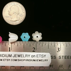 May include: Four plastic flower-shaped beads, one pink, one white, one blue, and one white with a hole in the center. The beads are lined up on a ruler, with the pink bead at the 1 inch mark, the white bead at the 1.5 inch mark, the blue bead at the 1.75 inch mark, and the white bead with a hole at the 2 inch mark. The ruler is labeled "IRIDIUM JEWELRY on ETSY WWW.ETSY.COM/SHOP/IRIDIUMJEWELRY SS STEEL".