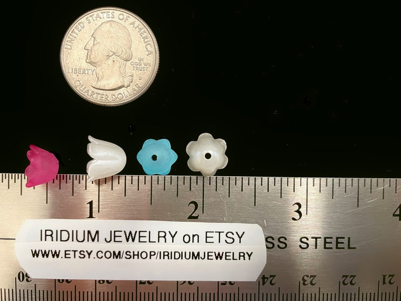 May include: Four plastic flower-shaped beads, one pink, one white, one blue, and one white with a hole in the center. The beads are lined up on a ruler, with the pink bead at the 1 inch mark, the white bead at the 1.5 inch mark, the blue bead at the 1.75 inch mark, and the white bead with a hole at the 2 inch mark. The ruler is labeled "IRIDIUM JEWELRY on ETSY WWW.ETSY.COM/SHOP/IRIDIUMJEWELRY SS STEEL".