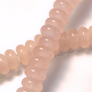 May include: Close-up of strands of light pink, oval-shaped beads. The beads are strung together, creating a necklace or bracelet. The beads have a smooth, polished surface and a translucent quality, with a soft, diffused light.