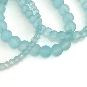 May include: Several strands of light blue frosted glass beads of varying sizes are arranged against a white background. The beads have a matte finish and are strung together, creating a delicate and ethereal appearance. Ideal for jewelry making.