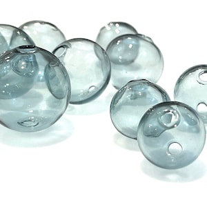 Hollow gray blue glass beads sit on a flat white surface.  These lampwork bubble beads have two holes and are shown in sizes  of 14mm 16mm and 20mm in diameter.  These are grey with a light blue cast.