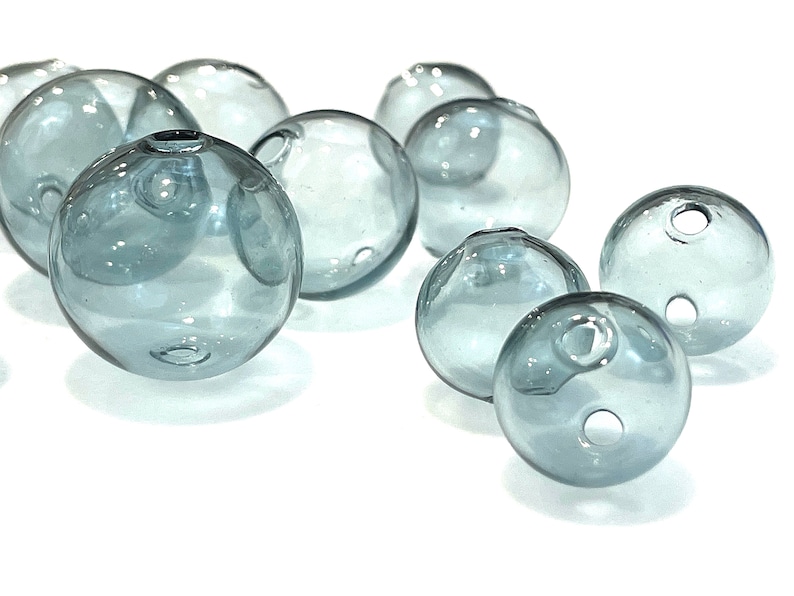 Hollow gray blue glass beads sit on a flat white surface.  These lampwork bubble beads have two holes and are shown in sizes  of 14mm 16mm and 20mm in diameter.  These are grey with a light blue cast.
