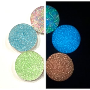 May include: Four round containers filled with iridescent glitter. The containers are arranged in a row. The glitter glows in the dark, with each container showing a different color: light blue, green, pink, and brown.