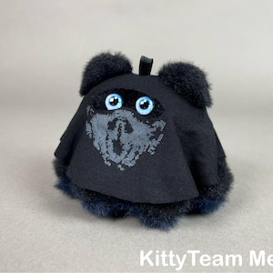May include: A plush, black creature with blue eyes and a gray face design. The creature has fluffy accents and a small loop at the top. The text "KittyTeam Meet" is at the bottom.