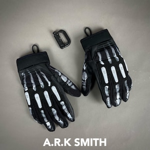 Puede incluir: Guantes negros con un diseño de huesos de esqueleto blancos. Los guantes tienen un puño negro y un lazo para colgar. También hay un mosquetón negro. El texto "A.R.K SMITH" está en la parte inferior.