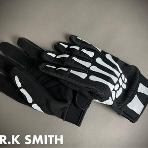 Op de afbeelding: Een paar zwarte handschoenen met een wit skeletbotontwerp op de achterkant van de hand. De handschoenen hebben een geribbelde textuur en de tekst "A.R.K SMITH" staat onderaan.