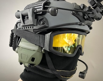 Pode incluir: Um capacete tático cinza com acessórios, incluindo óculos com lentes amarelas, um fone de ouvido e uma balaclava preta. O capacete tem um patch listrado em preto e branco. O texto "A.R.K SMITH" é visível na parte inferior.