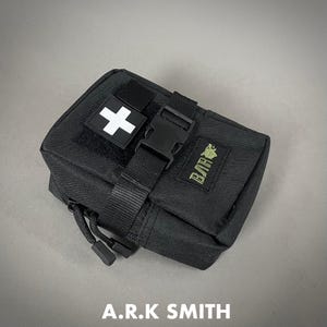 Op de afbeelding: Zwarte EHBO-kit met een wit kruispatch en een gespsluiting. De kit heeft een rits en een kleine groene patch met tekst. De tekst "A.R.K SMITH" staat onderaan afgedrukt.