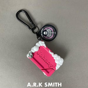 May include: A pink keychain with the name "Johnny" in a stylized font. The keychain has a black carabiner clip and a round black tag with a skull and wings design. White foam-like accents and a skull adorn the edges. The text "A.R.K SMITH" is at the bottom.