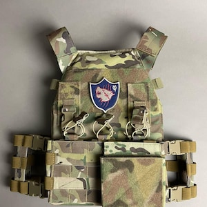 Puede incluir: Chaleco táctico de camuflaje con múltiples bolsillos, correas y hebillas. Cuenta con un parche con un emblema de lobo sobre un escudo azul. El texto "A.R.K SMITH" está impreso en la parte inferior. El chaleco está diseñado para uso militar o táctico.