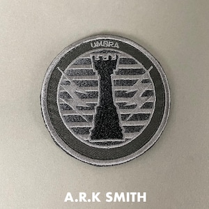 May include: Round patch with a black and grey design. The patch features a black rook chess piece in the centre, with the word "UMBRA" at the top. The text "A.R.K SMITH" is at the bottom.