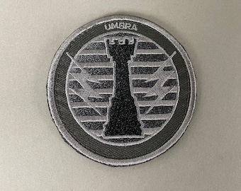 May include: Circular patch with a black and gray design. The patch features a black rook chess piece in the center, with the word "UMBRA" at the top. The text "A.R.K SMITH" is at the bottom.