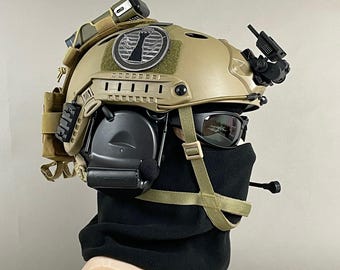 Pode incluir: Capacete tático bege com acessórios anexados, incluindo uma luz, fone de ouvido e dispositivo de visão noturna. O capacete apresenta um patch com um logotipo e é usado com um protetor de pescoço preto e óculos de sol. O texto "A.R.K SMITH" é visível na parte inferior.